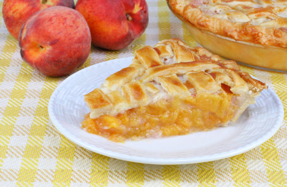 A slice of peach pie on a white plate on a white and yellow checked tablecloth