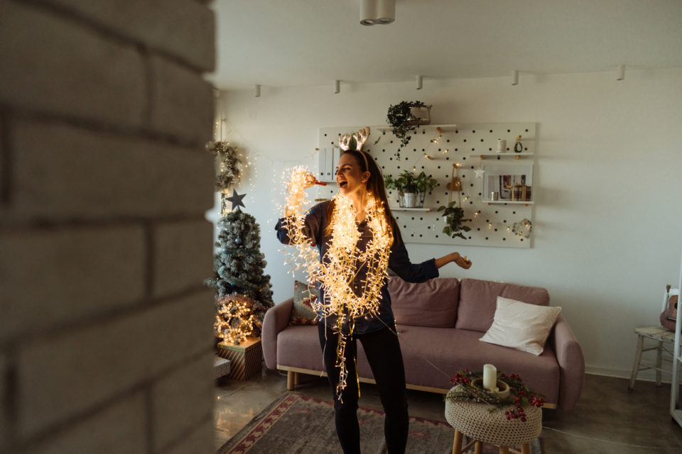 Photo of an excited young woman singing while decorating the living room of her apartment; wrapped in Christmas lights and wearing shiny reindeer antlers; preparing for the upcoming holidays.