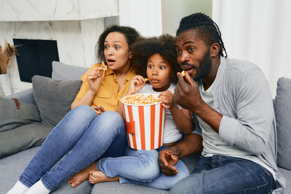 Father, mother and child sit on sofa with frightened faces during watching scary movie at home theater