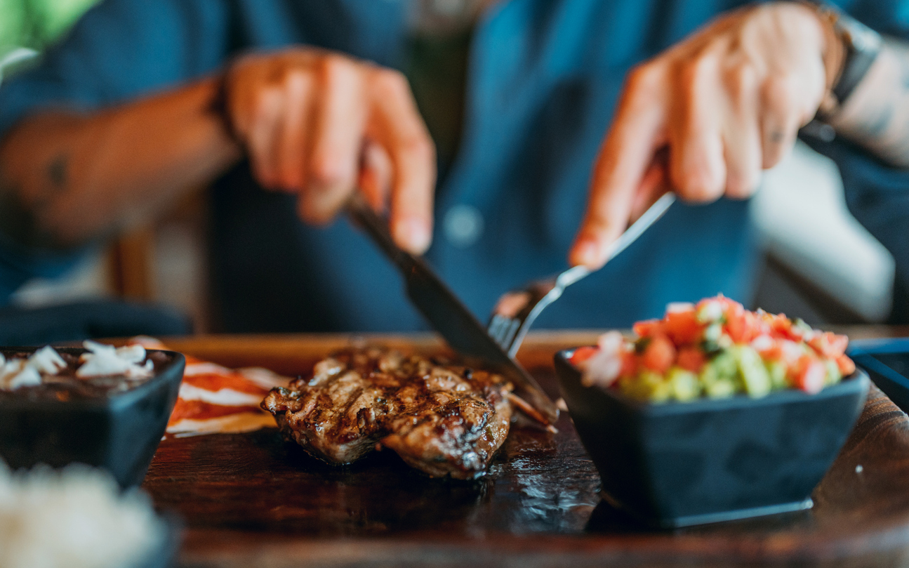Men's hands holding knife and fork, cutting grilled steak.