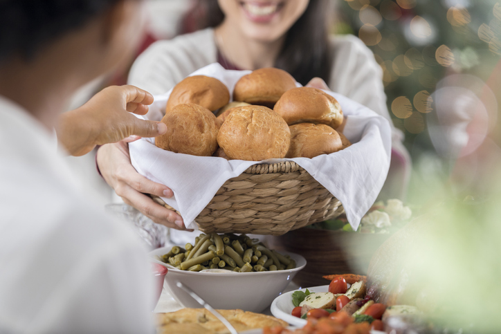 A smiling guest is unrecognizable as she reaches across a full Christmas dinner table with the bread basket to offer it to her unrecognizable friend. The friend reaches for a dinner roll from the basket.